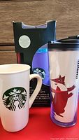 Photo showing white ceramic Starbucks mug, two Starbucks plastic tumblers, one with red fox design and one in original box