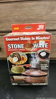 Front view of unopened Stone Wave Microwave Cooker box showing product name, images of prepared food, and product features.