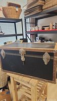 Closed black trunk chest showing metal reinforced corners, metal clasps and central lock with keyhole.