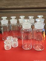 Front view of seven vintage clear glass bottles with glass stoppers, plus three extra stoppers in front on a red surface.