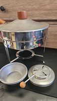 Vintage chafing dish with conical stainless steel lid with wooden knob and matching stand, fuel holder with ventilated cover, and round stainless steel serving pan with wooden handle.
