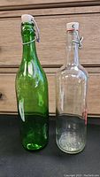 Two vintage glass bottles, one green and one clear, on a black surface against a wooden background, showing full side view of bottles including tops with swing stoppers.