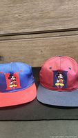 Two vintage Mickey Mouse snapback caps placed side by side on a dark surface with wooden drawers in the background. One cap is blue with red bill, the other red with gray bill. Both have large embroidered letter 'H' and Mickey Mouse on front.