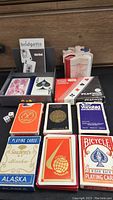 Photo showing multiple decks of vintage playing cards including Alaska, Bridgette, Piatnik, Yonkers Raceway, Maxicana, Vanolili. White and red casino chips and two dice also visible.