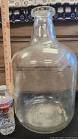 Front view of the large vintage Pyrex glass jug beside a water bottle for scale. Shows clear glass with scratches and marks inside and outside.
