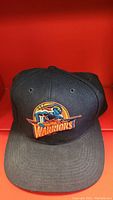 Front view of the black baseball cap showing the Golden State Warriors logo embroidered in orange, blue, and yellow.