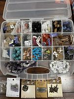 Top view of a plastic organizer box containing multiple compartments with various beads in different colors and materials such as turquoise, black, white skull shapes, amber hues, and metallic findings. Some compartments contain packaged charms and findings.