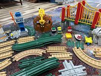 Wide assortment of interlocking green Geotrax track pieces, four building structures including yellow-red bridge and mountain, and various toy vehicles and boats arranged in a display.