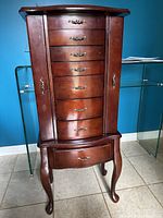 Front view of wooden jewelry armoire with seven drawers and closed side doors, showing dark wood finish and curved legs