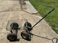 Metal utility cart with green frame and wire basket, four black rubber air-filled wheels, and long handle with plastic grip, positioned on concrete outdoors.