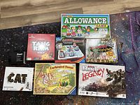 Photo showing 7 boxed board games: Allowance, Tangram Race (sealed red box), Laser Maze with plastic board, Night at the Museum, The Cat Game, Enchanted Forest, Risk Legacy.