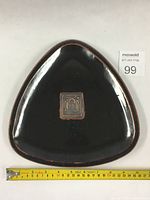 Top-down view of the triangular black pottery plate with a Buddhist stamp in the center and measuring tape showing approximately 9 inches per side.