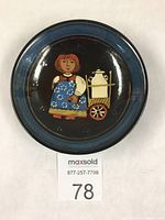 Top view of the round pottery dessert plate showing detailed folk art painting of a person with a cart and kettle in bright mid-century colors on a dark background with a blue border.
