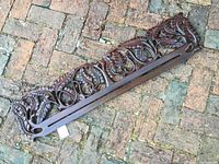 Top view of one carved teak ikat hanger resting on a brick surface, showing detailed leaf and fern carving and central slit.
