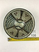 Top view of a circular pottery bowl signed by Meg Burgess, showing the speckled glaze, brown radials, blue crisscross design, and central blue curves.