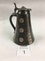 Front and side view of the salt glazed syrup jug showing cobalt blue and iron oxide colors with raised floral designs and a metal lid.