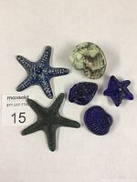 Top view of six decorative seashell and starfish items on white background, showing variety of cobalt blue ceramic/glass pieces and a real conical shell.