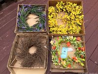 Four faux floral wreaths in their original storage boxes, including a twig wreath, yellow flower wreath, lavender wreath, and a colorful spring wreath with orange flowers.