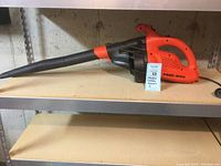 Full side view of the orange and black electric blower on a shelf showing the entire length with nozzle attached and the power cord.