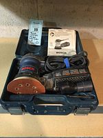 Full kit including Bosch 1295DVS sander, blue plastic carrying case, sanding pad attached, vacuum attachment, power cord, and instruction manual inside case.