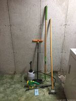 Full set of garden tools including shovel, rake, hoe, sprayer, and feeder/waterer against concrete wall and green carpet
