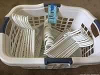 White plastic laundry basket filled with 50 white plastic hangers, showing vent holes and dark blue handles.