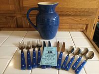 Blue ceramic pitcher with cream interior and 12 pieces of blue handled flatware consisting of forks, knives, and spoons arranged on white tiled surface with wooden background.