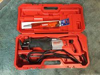 Open red hard case holding Milwaukee reciprocating saw, power cord coiled, and two spare blades in packaging.