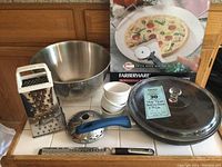 Wide shot showing all items in the lot arranged on a counter including mixing bowl, graters, ramekins, baking stone, and cutting tool