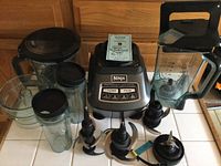 Full view of Ninja blender base and attachments including large blending jar, two smaller blending cups, multiple blade attachments laid out on a countertop.
