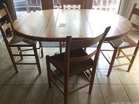 Oak kitchen table with four chairs arranged around it. Table is oval with leaf inserted and shows wood grain and finish.