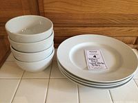 Stack of 4 white ceramic bowls next to stack of 4 matching dinner plates with label indicating Pottery Barn Great White Pottery.