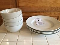 Four white bowls stacked on the left and four white dinner plates stacked on the right against wooden cabinetry.