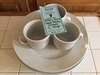 Three white ceramic mugs stacked inside and placed on top of stacked white plates, with a lot tag.