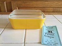 Front angle of yellow Pyrex fridgie with glass lid and auction label.