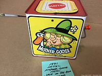 Front view of vintage 1971 Mattel metal Jack-in-the-Box, featuring Mother Goose illustration and red crank, in good condition.