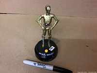 Front view of a gold-colored C-3PO talking statue standing on black base with 'Star Wars' logo and yellow button. A black and white Sharpie marker is placed next to it for scale.
