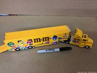 Full side view of diecast yellow M&M's Racing Team semi truck and trailer with colored M&M characters and logos, Sharpie pen placed beside for scale.