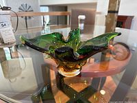 Front angle photo of the green hand blown glass ashtray on a glass table showing its organic leaf-like form and colors.