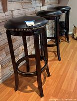 Image showing three bar stools with dark wooden bases and black faux leather seats against a stone wall and wood floor.