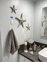Bathroom corner with three starfish wall art pieces in varying sizes, gray waffle towel on hook, round table mirror, soap dispenser, and small hexagonal mirror tray on dark countertop next to white sink.