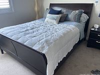 View of queen size bed made up with white quilted duvet, pillows, and sheets on a dark wooden bed frame, in a bedroom with carpeting and window with white blinds.