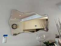 Front view of the wall mirror mounted on a light-colored wall. Reflective surface shows part of a living area with ceiling lights and wall clock.