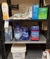 Shelving unit with various cleaning and laundry supplies including bottles of dishwashing liquid, Olay body wash, Nellie's laundry soda, and boxes of napkins and Swiffer cloths.