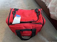Red emergency bag with black straps and multiple compartments, labeled 'Emergency Bag'. A blue and white tag is attached.