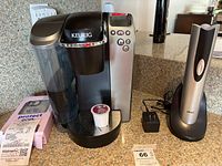 Keurig coffee maker with a single K-cup inserted, Oster electronic pepper mill on charging base, power adapter, and iPhone 12/12 Pro cover box grouped on kitchen countertop.