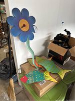 Image of blue flower shaped clock with visible clock hands and green stem near a broom and boxes