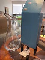 Clear glass art vase next to original blue Nambe box on a table with wood and glass surface.