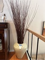 Full view of large beige ceramic vase filled with tall dried willow branches, positioned on floor next to wooden cabinet and stair railing.