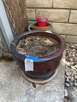Large reddish-brown plant pot with soil and dried leaves inside, resting on a matching saucer, outdoor setting.